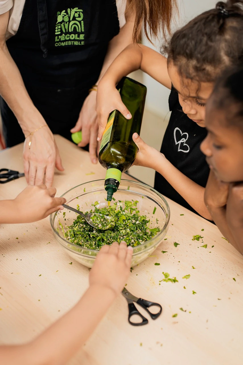 Trois écoliers accompagnés d'une professeur sont installés sur une table préparent une salade fraiche en versant de l'huile d'olive dans un saladier